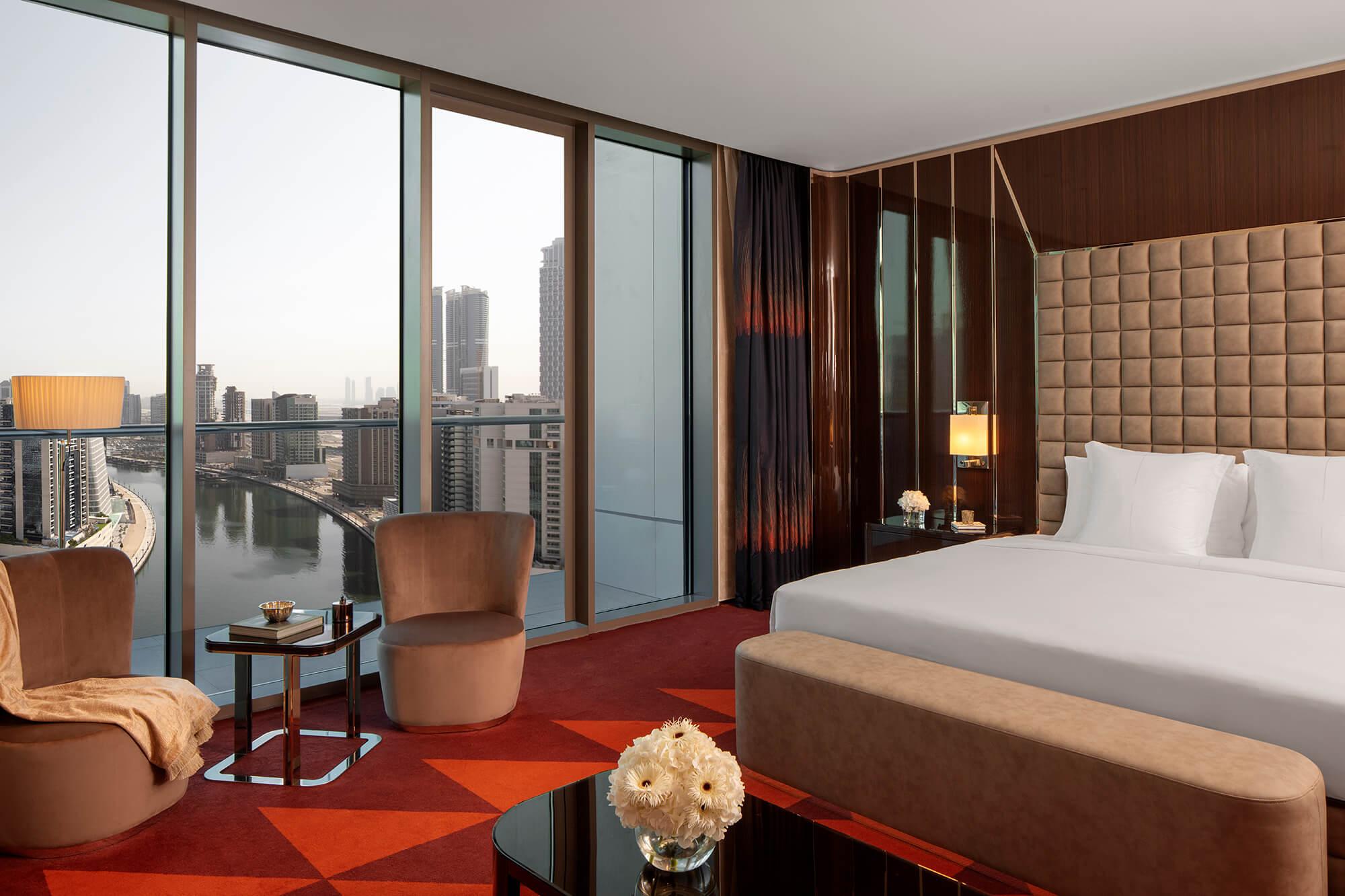 A king size bed with bedroom bench on the right, with a coffee table in the foreground, 2 armchairs and coffee table to the left next to a top to bottom window displaying high rise views of Dubai city and canals