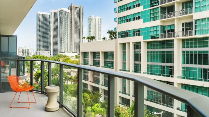 Outdoor balcony with orange chair, small beige table, looking out to another building with turquoise windows, and other high-rises in the distance, with palm trees scattered throughout