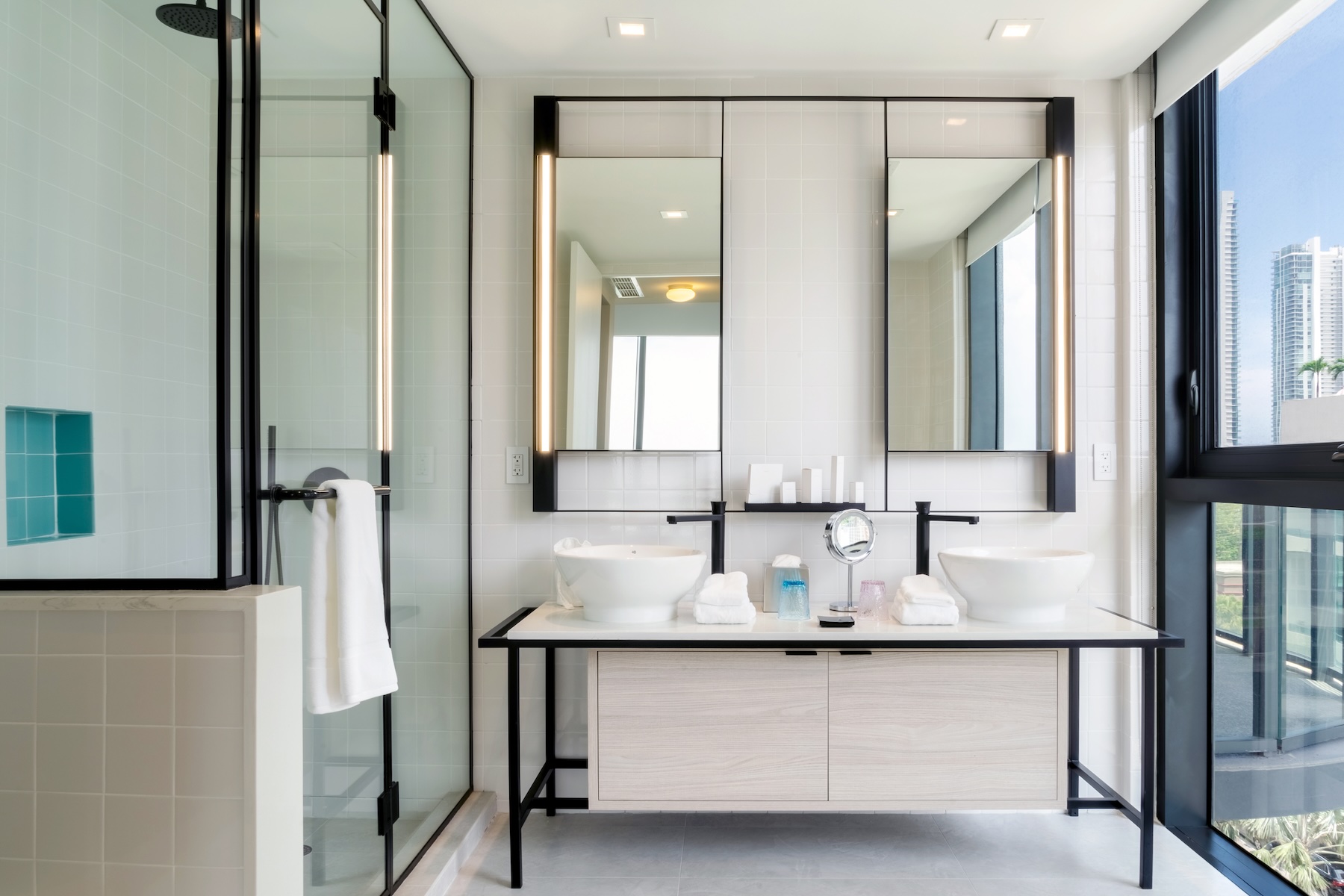 A modern bathroom with two sinks in the centre of the image, with walk in glassed shower on the left, and floor to ceiling window with city views