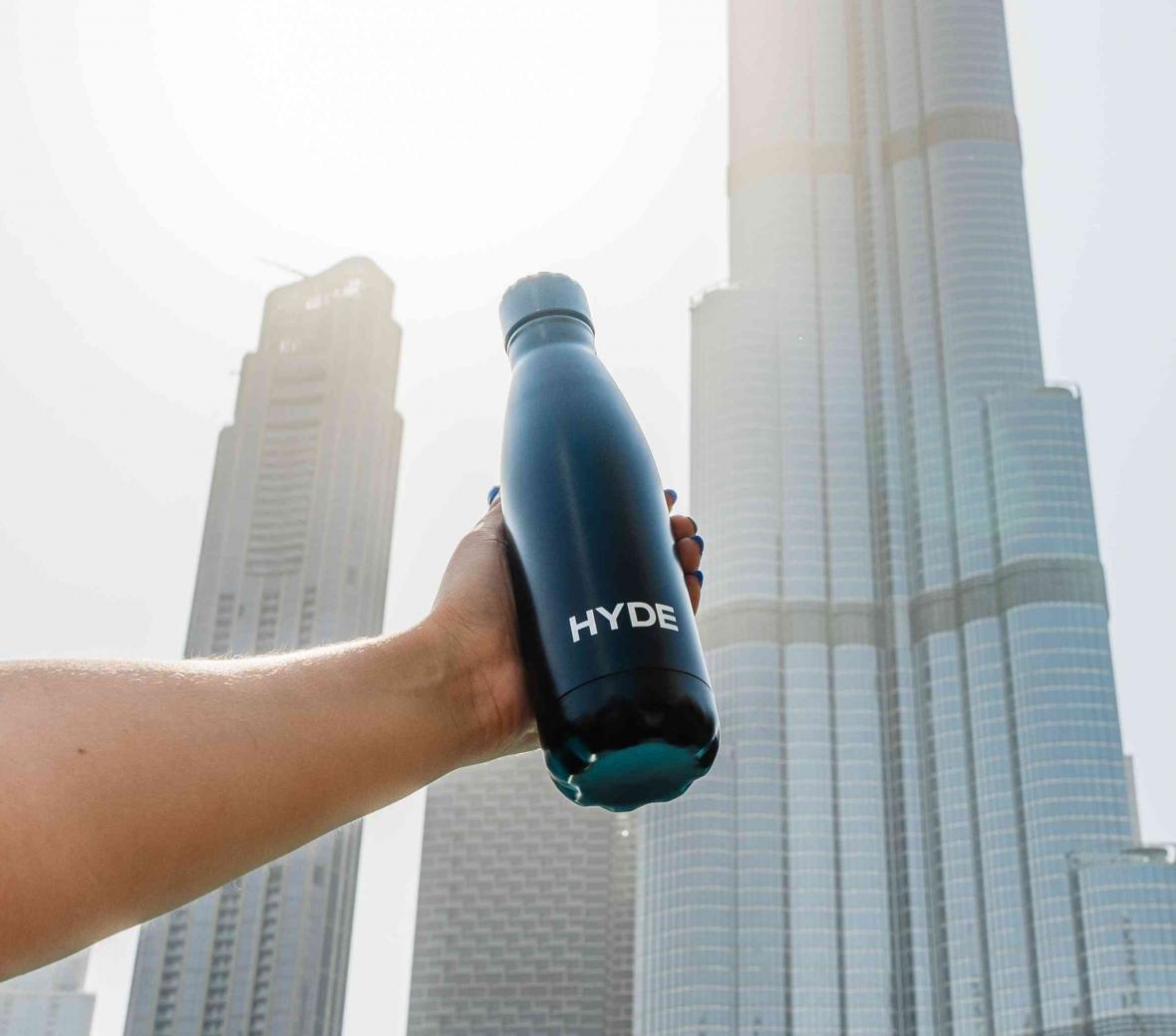 Black water bottle with HYDE written in white being held up by an arm with skyscrapers in the background