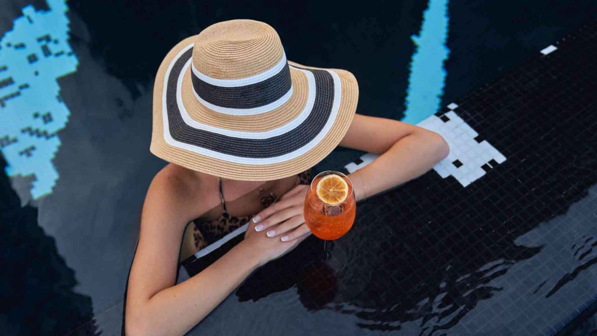 Woman in striped hat leaning against pool ledge with orange spritz in a glass
