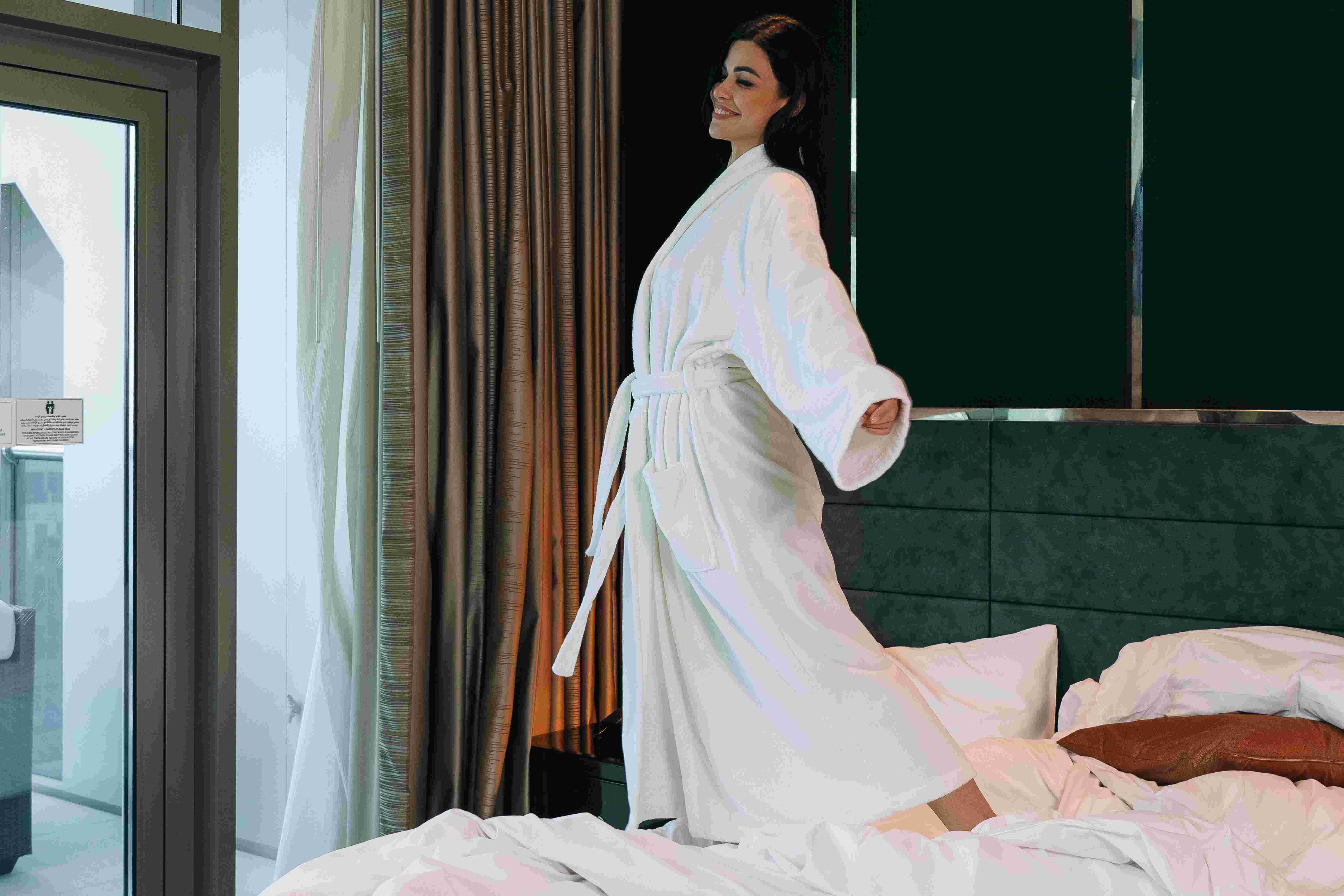 Woman in white robe standing on top of white bed with grey headboard and gold curtains in the background