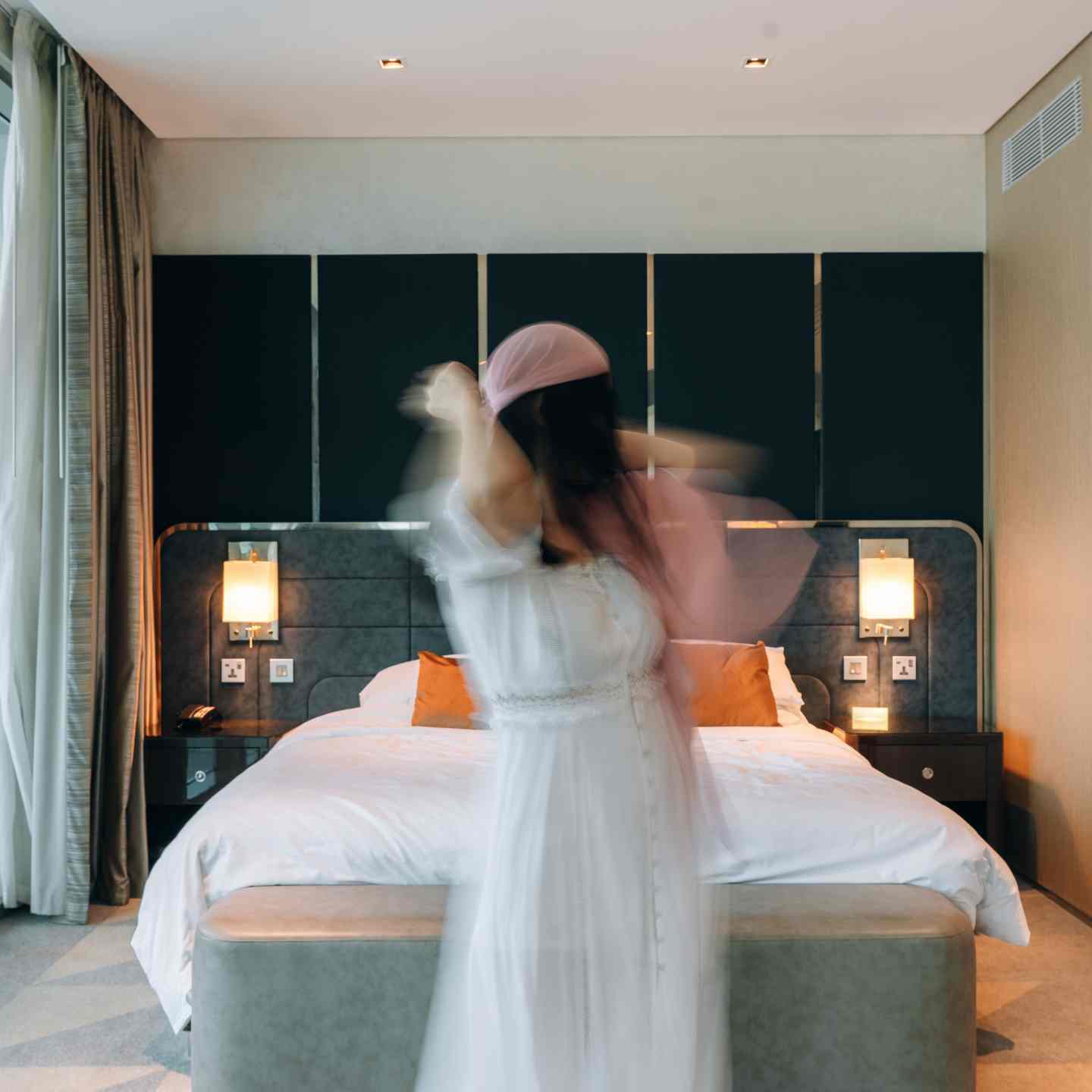 Woman in white dress and light pink headwrap dancing in front of white king bed with grey headboard