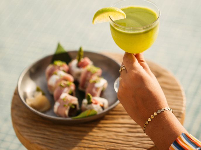 Green margarita being held by hand with sushi on a wooden stool in front of a pool in background