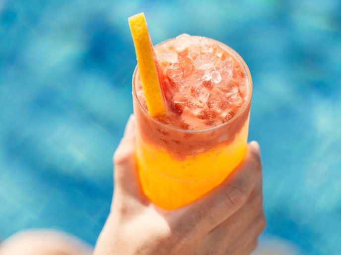 Orange cocktail with orange slice held by someone's hand with pool in the background