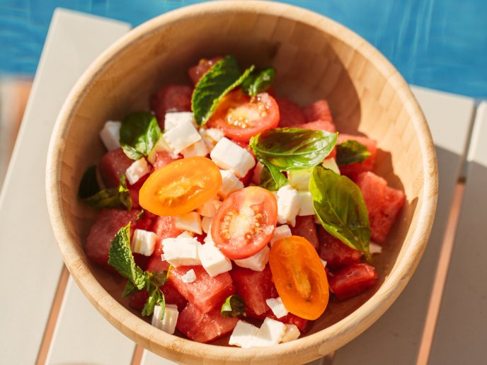 Tomatoes, cheese, and basil salad in a bowl with orange cocktail and pool in background