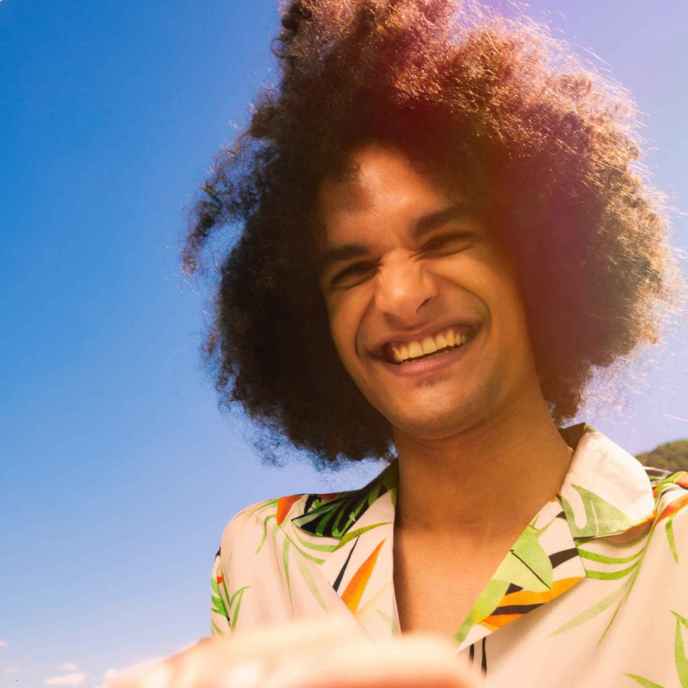 Man with curly hair smiling in collared shirt with blue sky in background