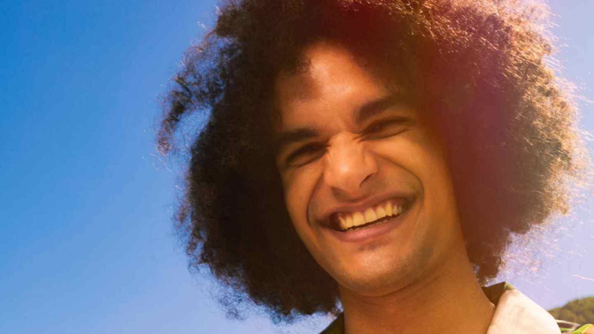 Man with curly hair smiling with blue sky in background