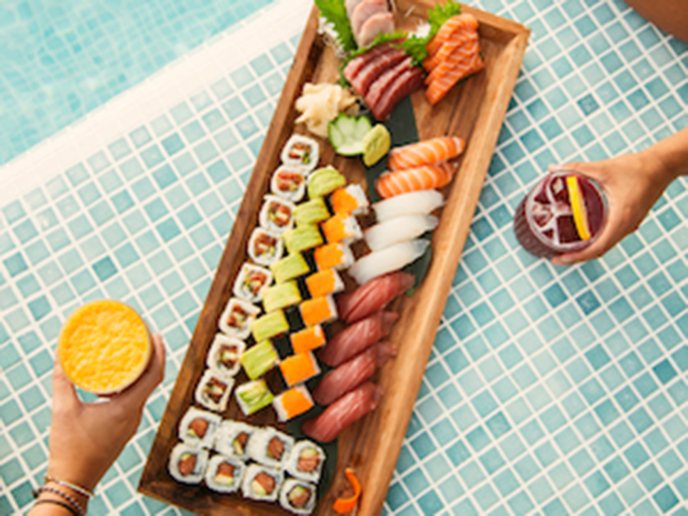 Sushi on a wooden tray on top of a pool with womens legs and arms in the background