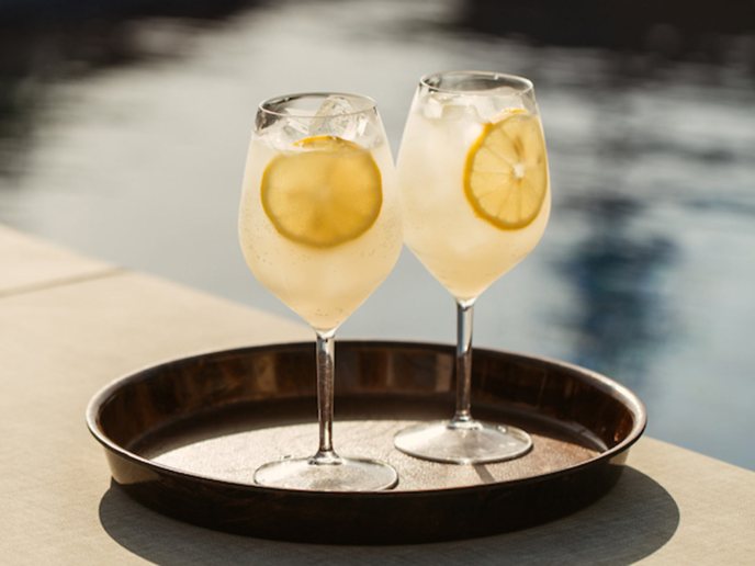 Two yellow cocktails on a tray with lemon slices in them with pool in background