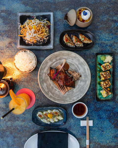 Meat, rice, sushi, and a cocktail sitting on various shaped plates on a gray table top