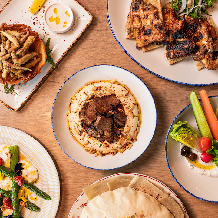 Hummus, pita, vegetables and more on white round and rectangular plates on a light wood table
