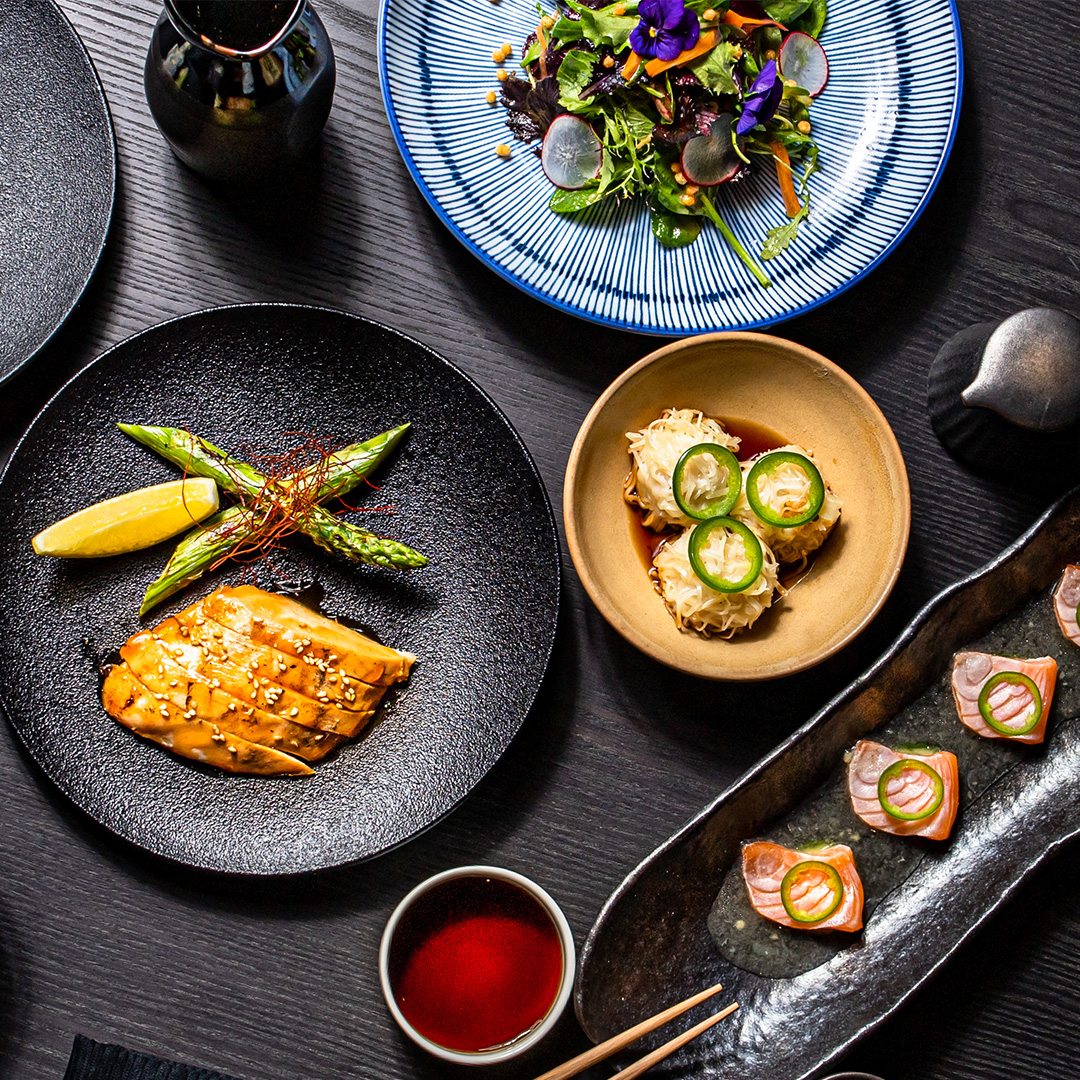 Chicken, asparagus, sushi, and more on various shaped plates on a black tabletop