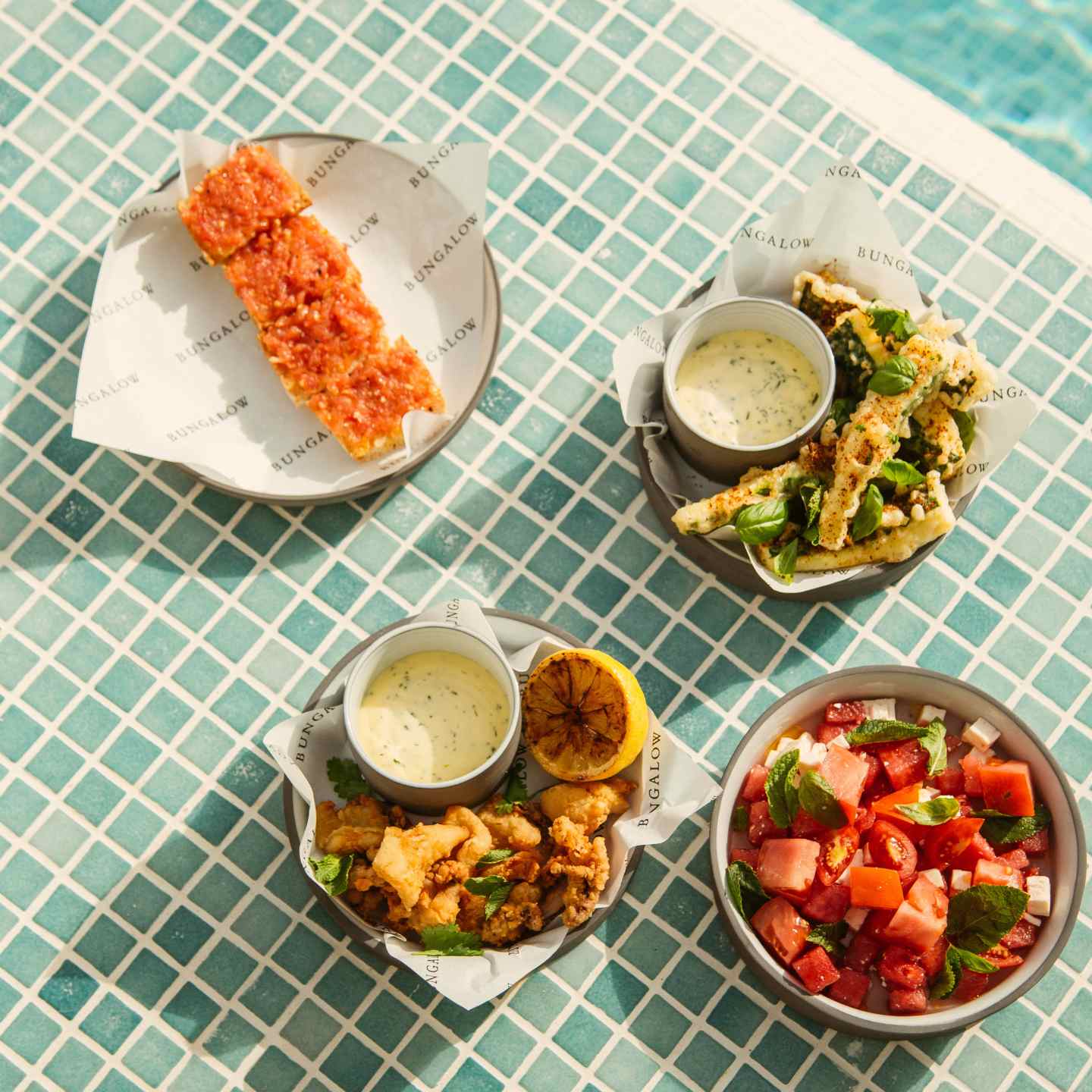Tomato salad in a round bowl, calamari with dipping sauce and lemon wedge, pan con tomato on round plate, fried zucchini with dip all on a blue pool tile