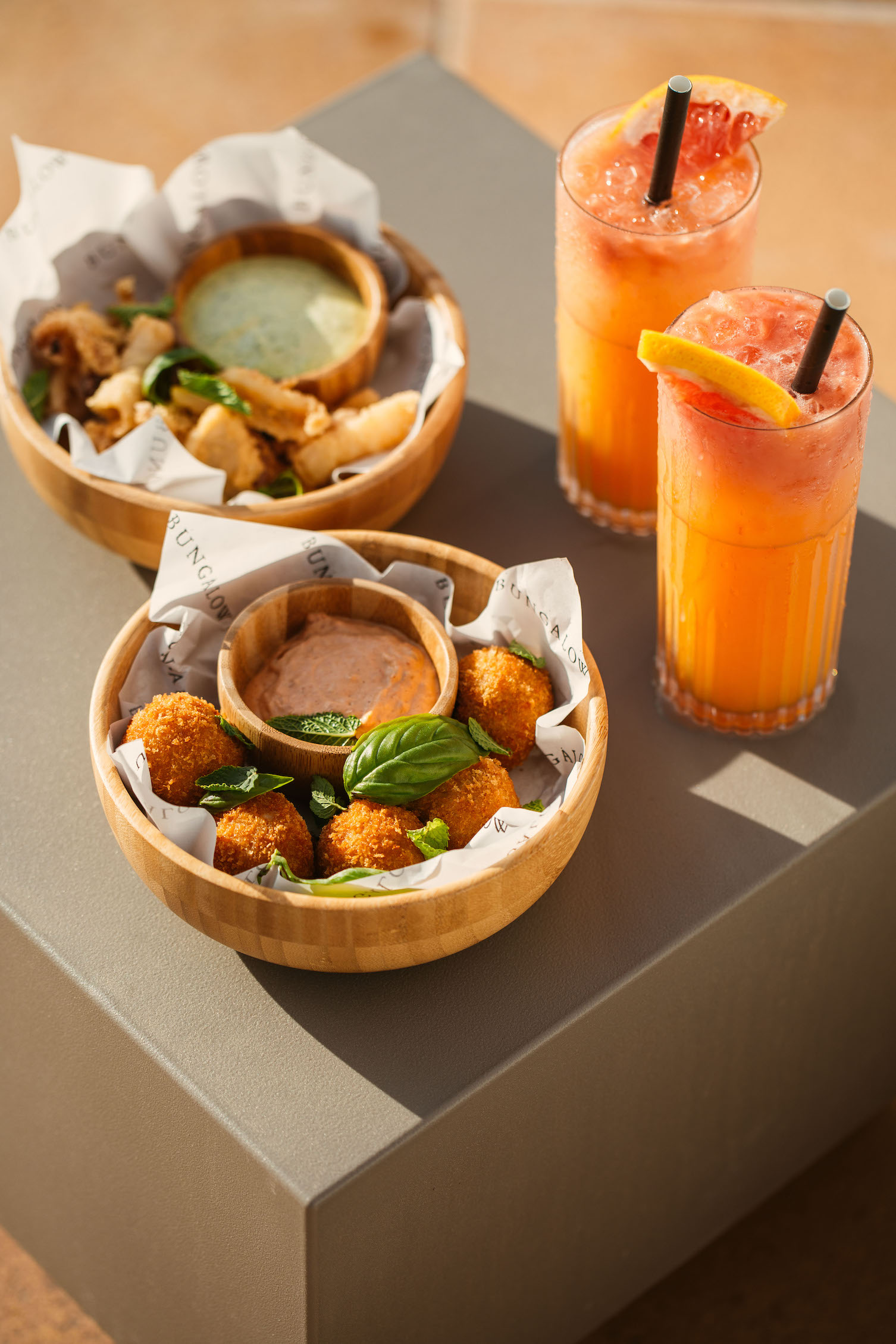 Appetizers with dipping sauces in wooden bowls with orange cocktails sitting on a grey square table