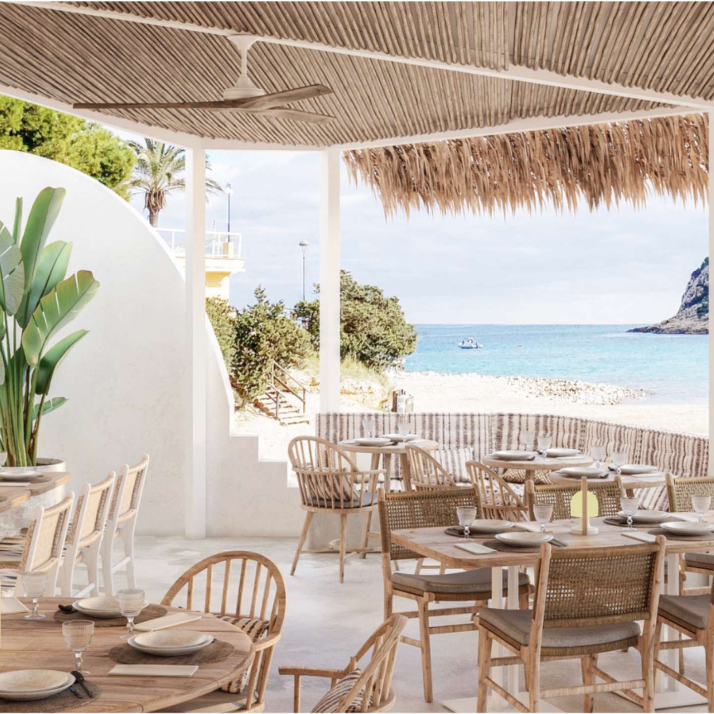 Outdoor restaurant space with light wooden and wicker chairs and tables with white walls and straw patio cover overlooking a beach and blue sea