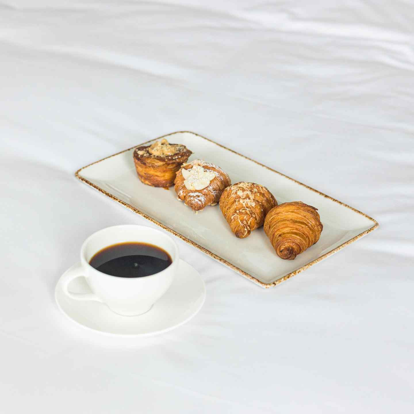Small cup of coffee in white coffee cup and four croissants on a rectangular plate sitting on a white bed