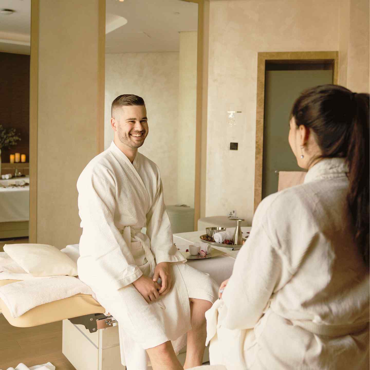 Man and woman in white robes sitting on massage tables smiling at each other