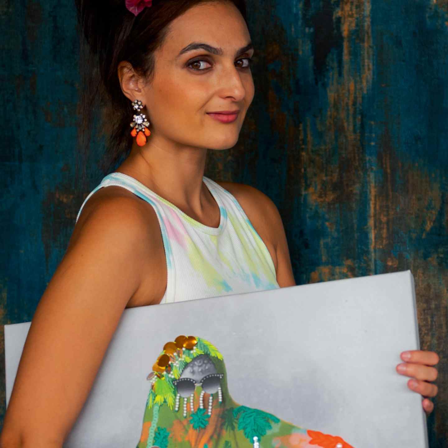 Woman with large elaborate earrings poses with a piece of artwork against a blue background