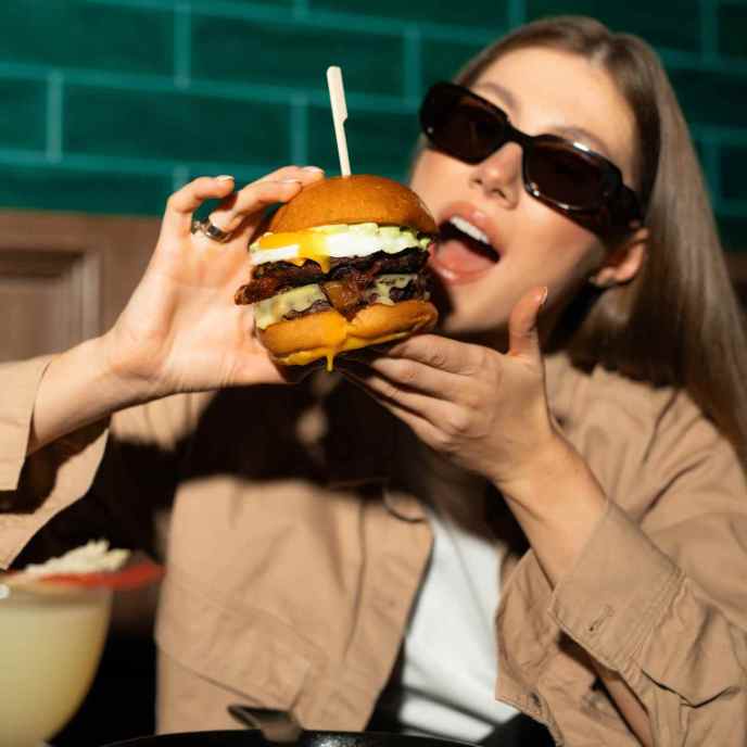 Girl with black sunglasses and tan blouse holds up a large cheeseburger and bites into it