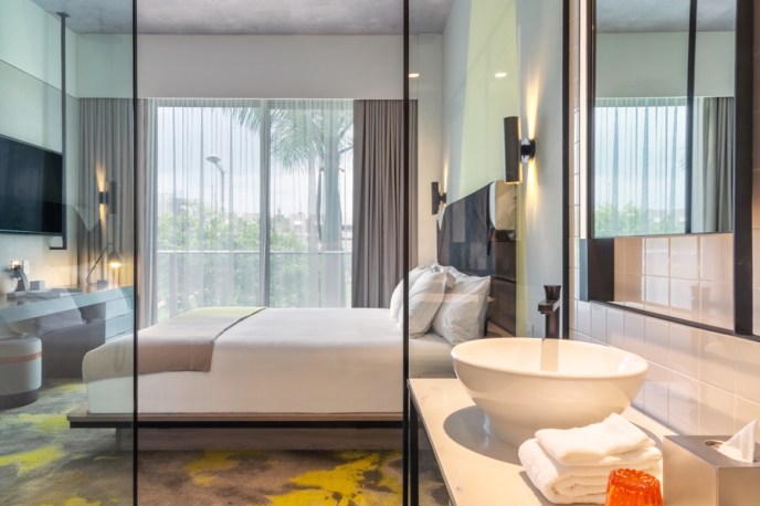 Hotel bedroom looking out from glass bathroom windows with white sink, large white bed, tv on wall, and large windows with grey curtains.