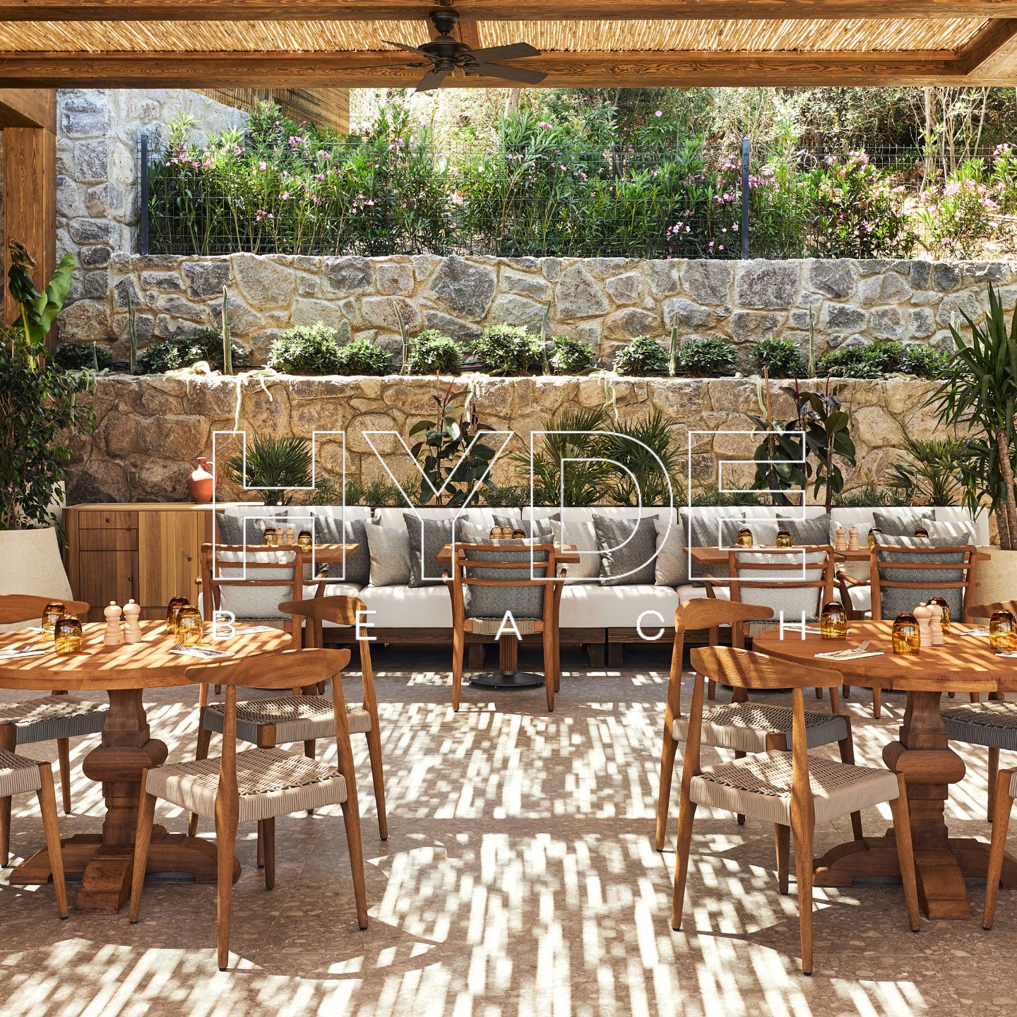 Outdoor restaurant with wooden tables and wooden chairs, stone wall, and patio overhead