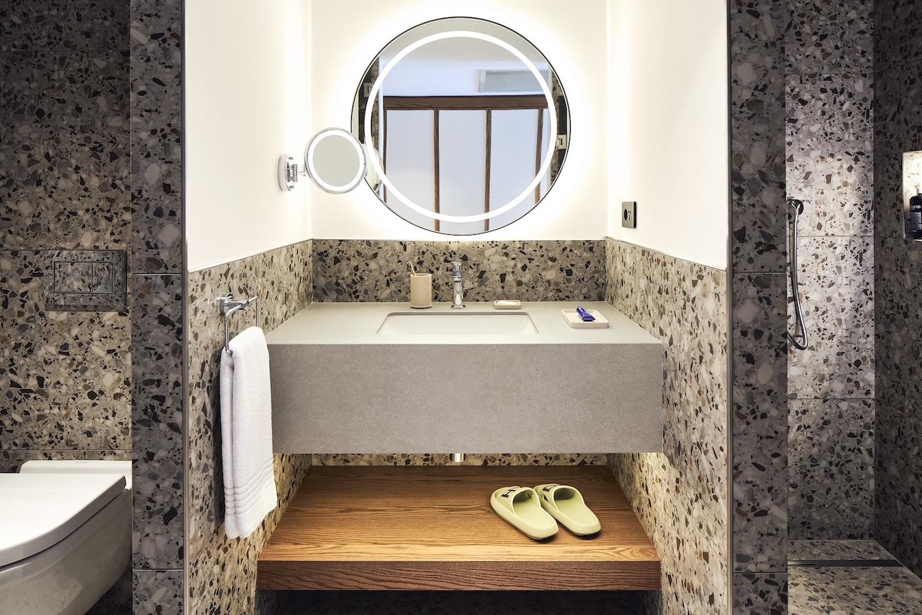 Grey bathroom sink with circular mirror and wooden shelf with sandals on it