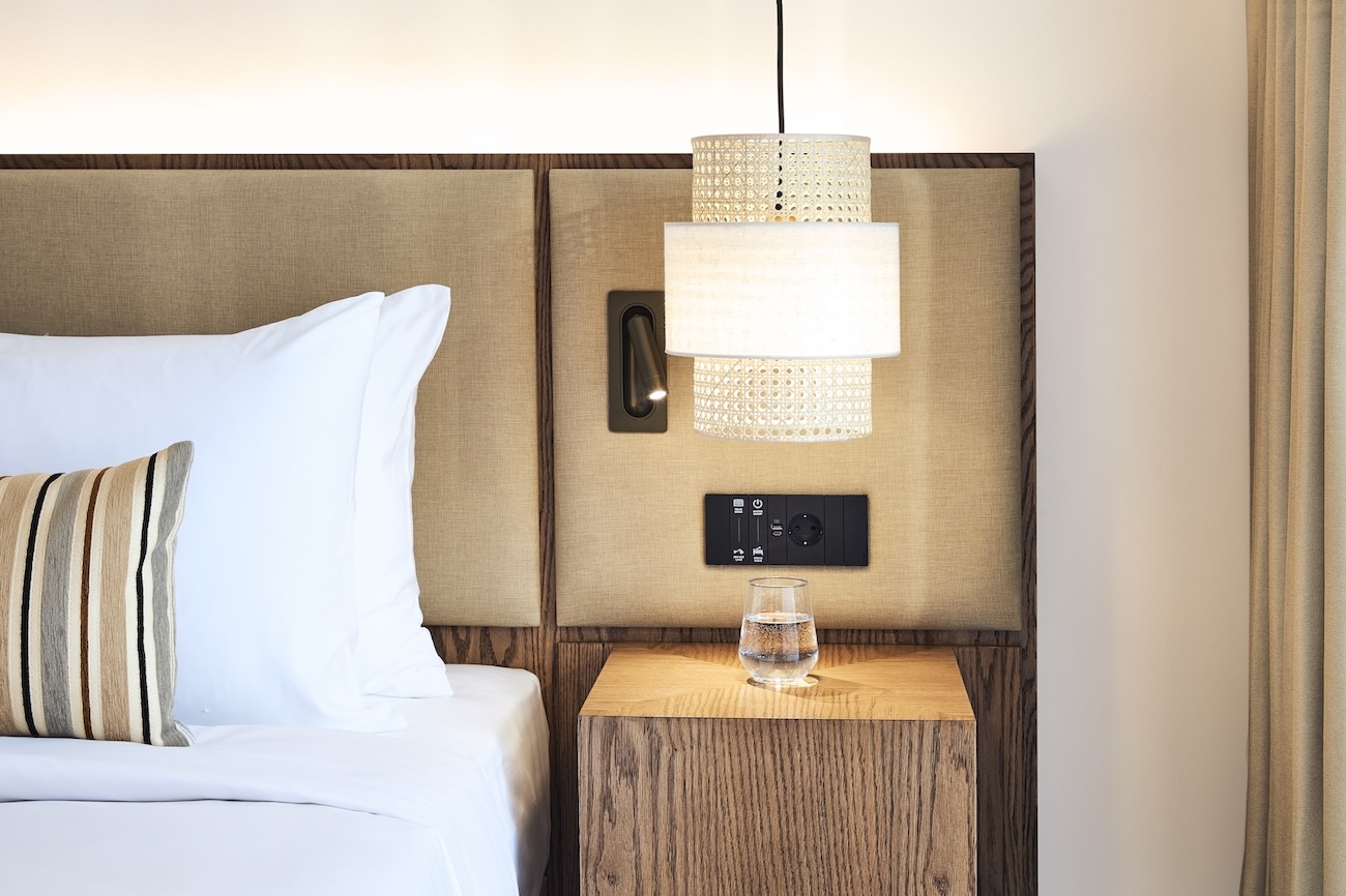 Corner of white bed with beige headboard and wooden bedside table with pendant light hanging