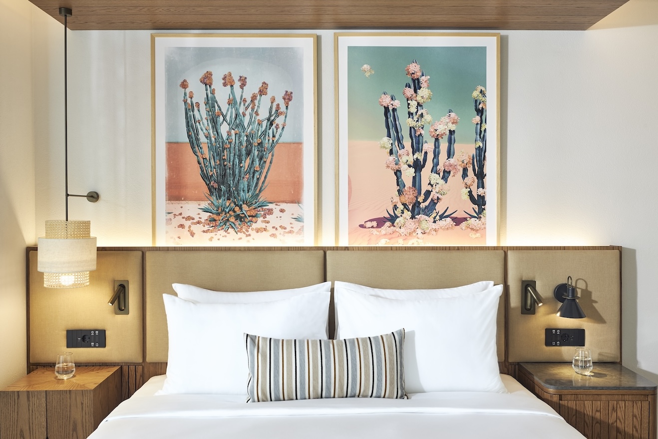 Straight on shot of large bed with beige headboard, striped pillow, two bedside tables, pendant light, and two framed paintings of flowers on the wall