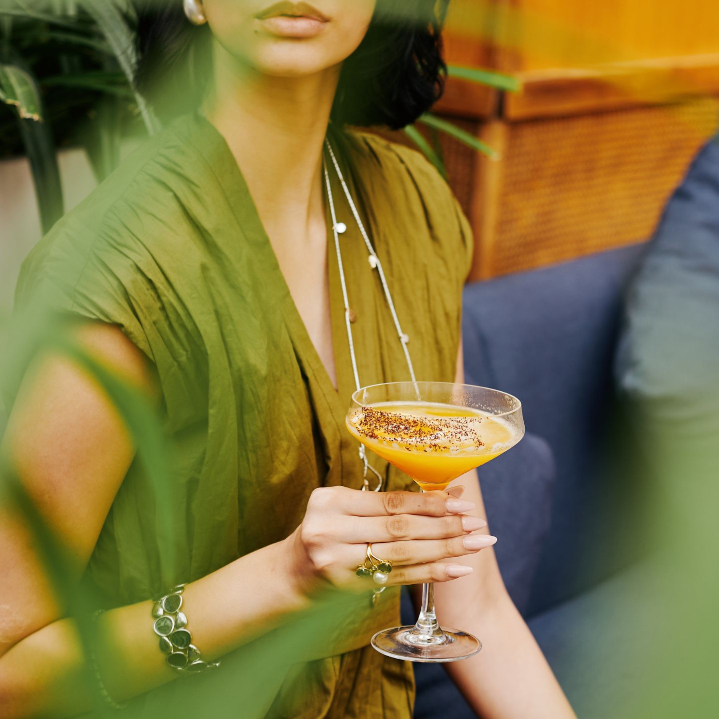 Woman in green top holds an orange cocktail in a coupe glass with greenery surrounding her