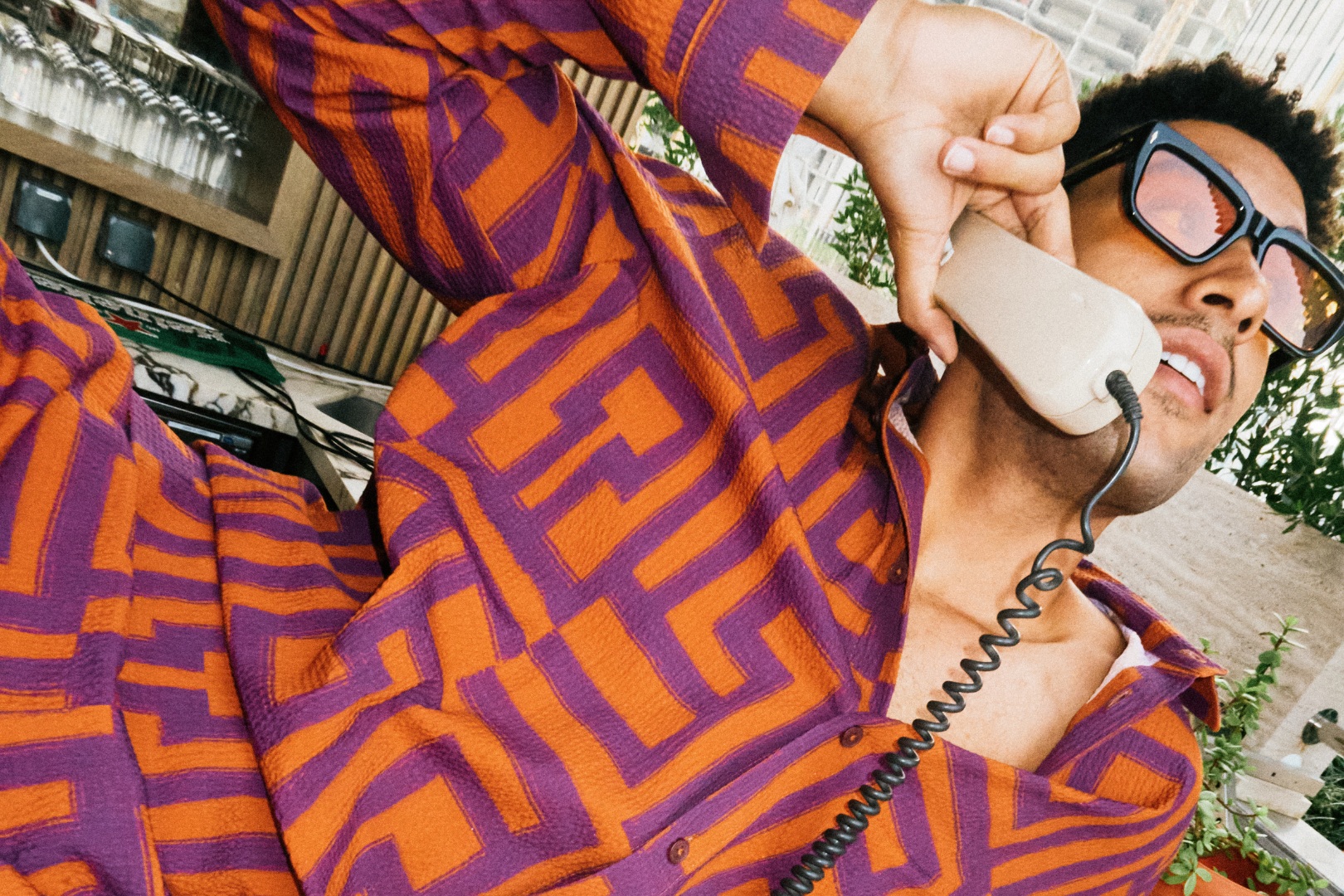 Man with curly hair in orange and purple patterned shirt puts a cord phone up to his ear
