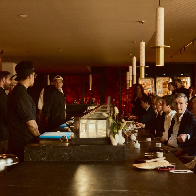 Guests sit at a dark sushi counter while sushi chefs stand behind the counter