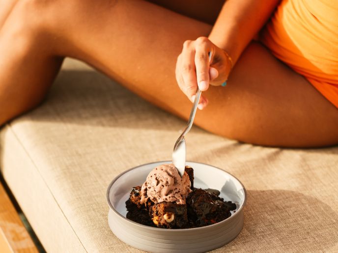 Brownie and chocolate ice cream in a bowl on a beige cushion with a woman's hand holding a spoon dipping into the ice cream