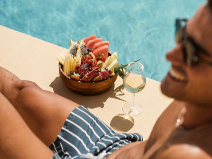 Man in striped swim trunks and sunglasses smiles with a bowl of fruit and glass of wine nearby a pool