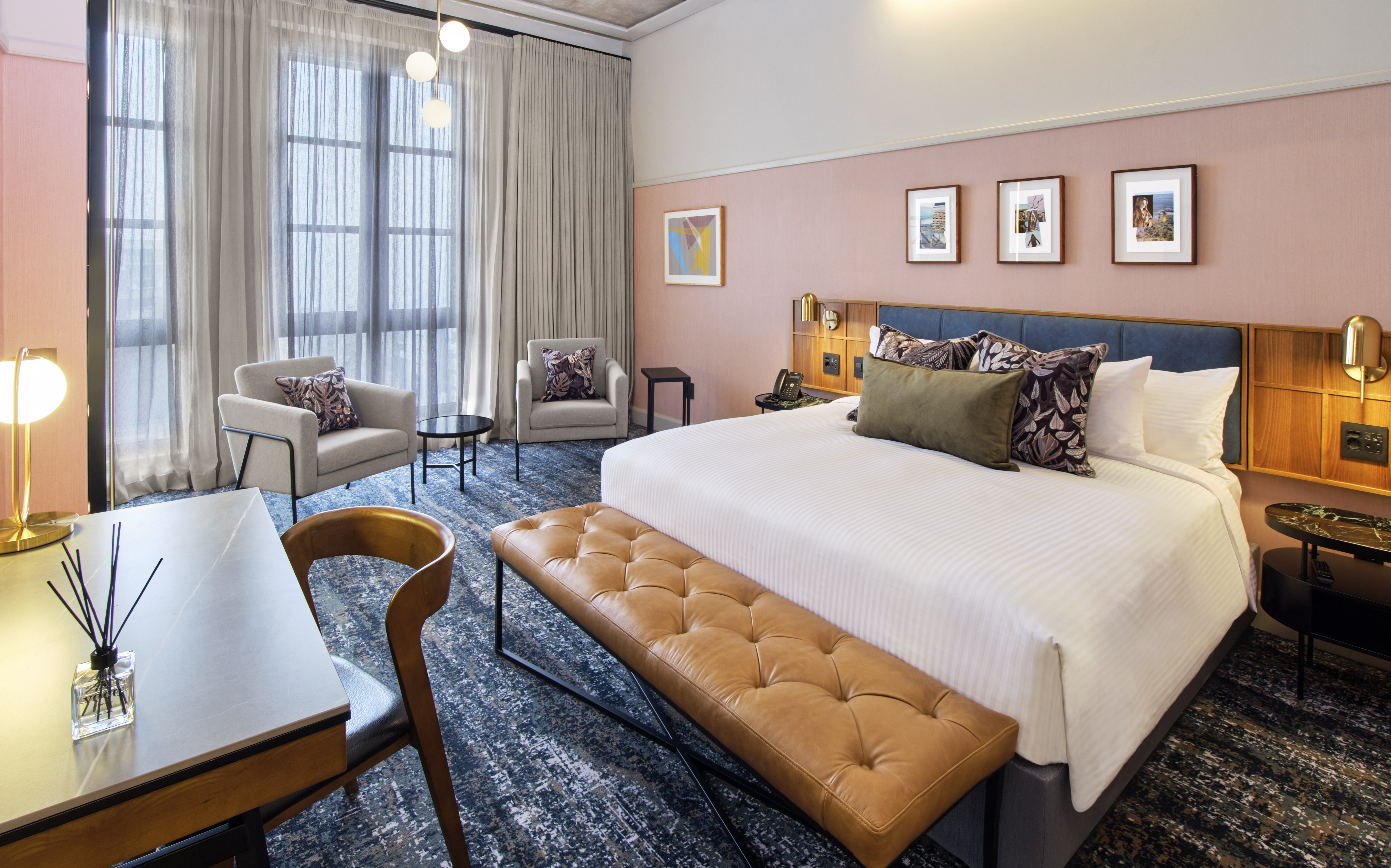 This is an image of a hotel room featuring a contemporary and stylish design. The room has a large bed with a tufted leather bench at the foot. The walls are decorated with soft pink tones and framed artwork above the headboard. The room includes a large, illuminated mirror with globe lights next to the bed, a wall-mounted TV, and a side table with a modern lamp and outlets. The window has sheer curtains, allowing natural light to filter in, and the ceiling shows an exposed concrete design.