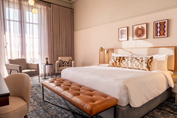 This is an image of a hotel room featuring a contemporary and stylish design. The room has a large bed with a tufted leather bench at the foot. The walls are decorated with soft pink tones and framed artwork above the headboard. The room includes a large, illuminated mirror with globe lights next to the bed, a wall-mounted TV, and a side table with a modern lamp and outlets. The window has sheer curtains, allowing natural light to filter in, and the ceiling shows an exposed concrete design.