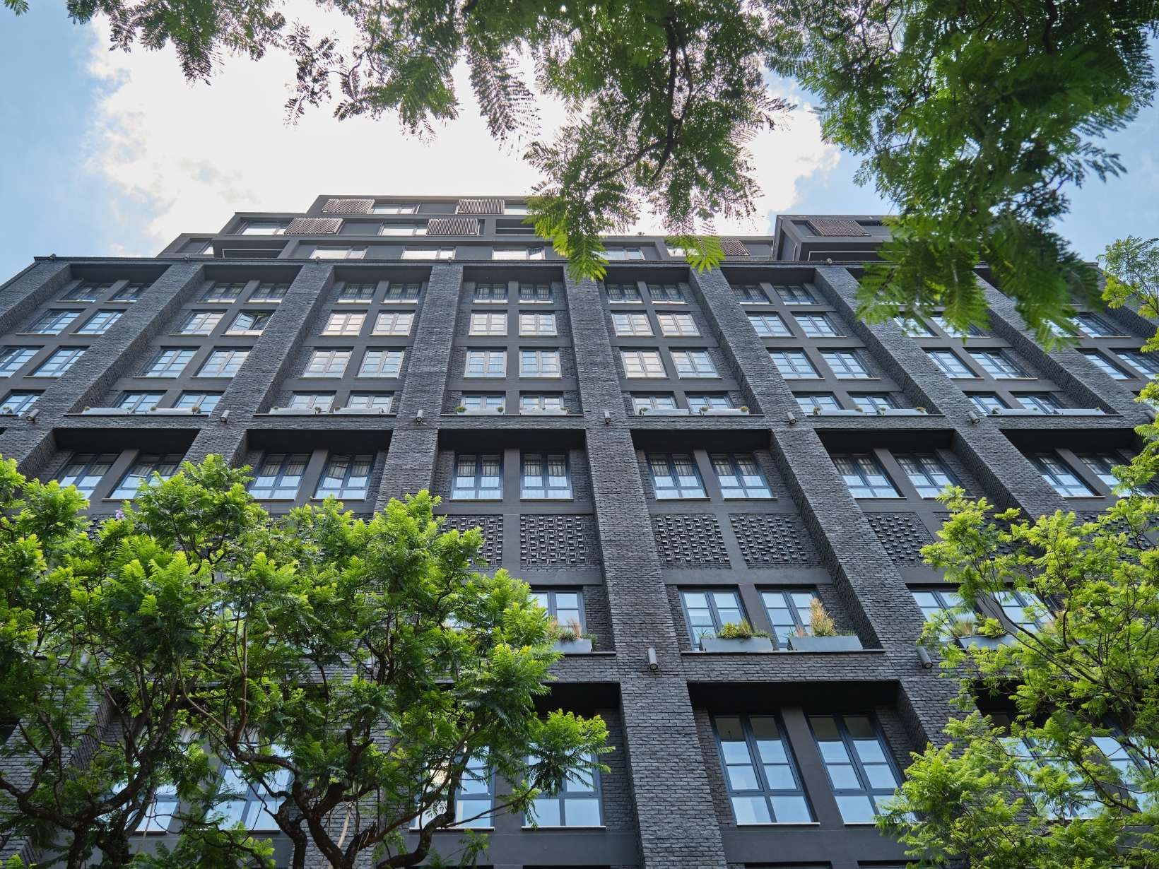 Grey building with large windows, blue sky, white clouds, and green trees in front of the building