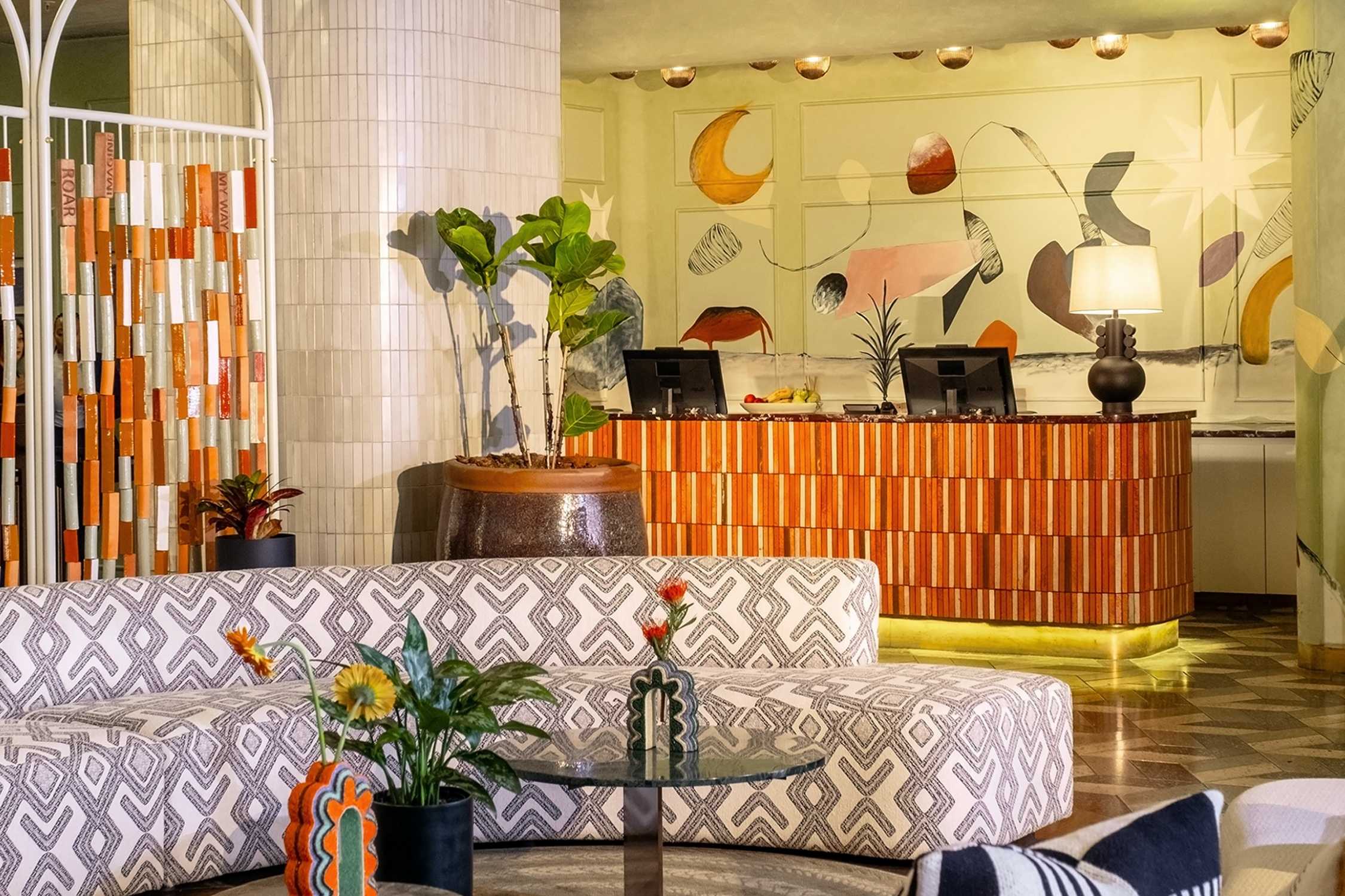 Warm tones at reception desk in front of the art wall, pillar with pot plants placed in front, and a white couch with black designs in Hyde Johannesburg.