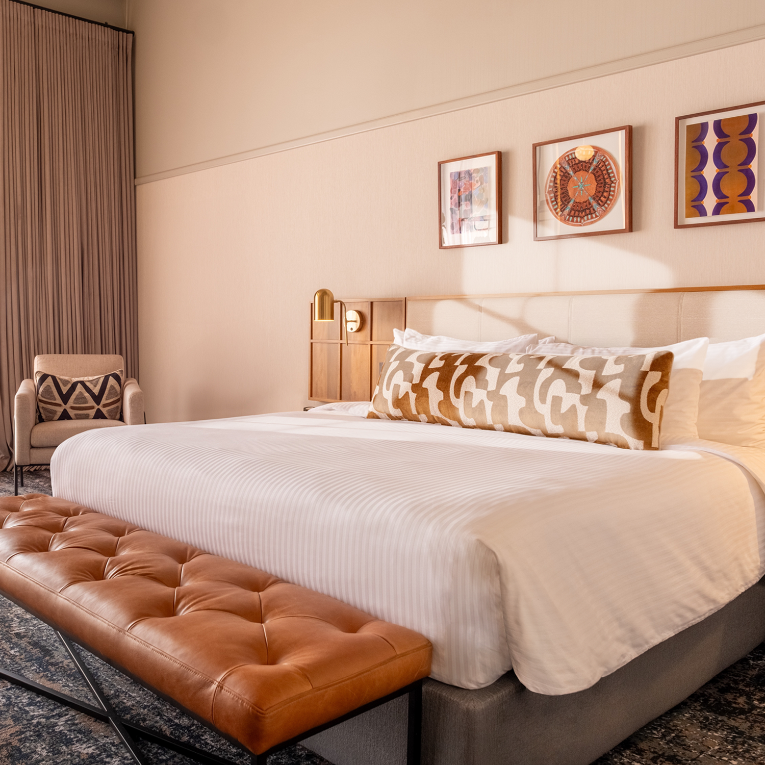 This is an image of a hotel room featuring a contemporary and stylish design. The room has a large bed with a tufted leather bench at the foot. The walls are decorated with soft pink tones and framed artwork above the headboard. The room includes a large, illuminated mirror with globe lights next to the bed, a wall-mounted TV, and a side table with a modern lamp and outlets. The window has sheer curtains, allowing natural light to filter in, and the ceiling shows an exposed concrete design.