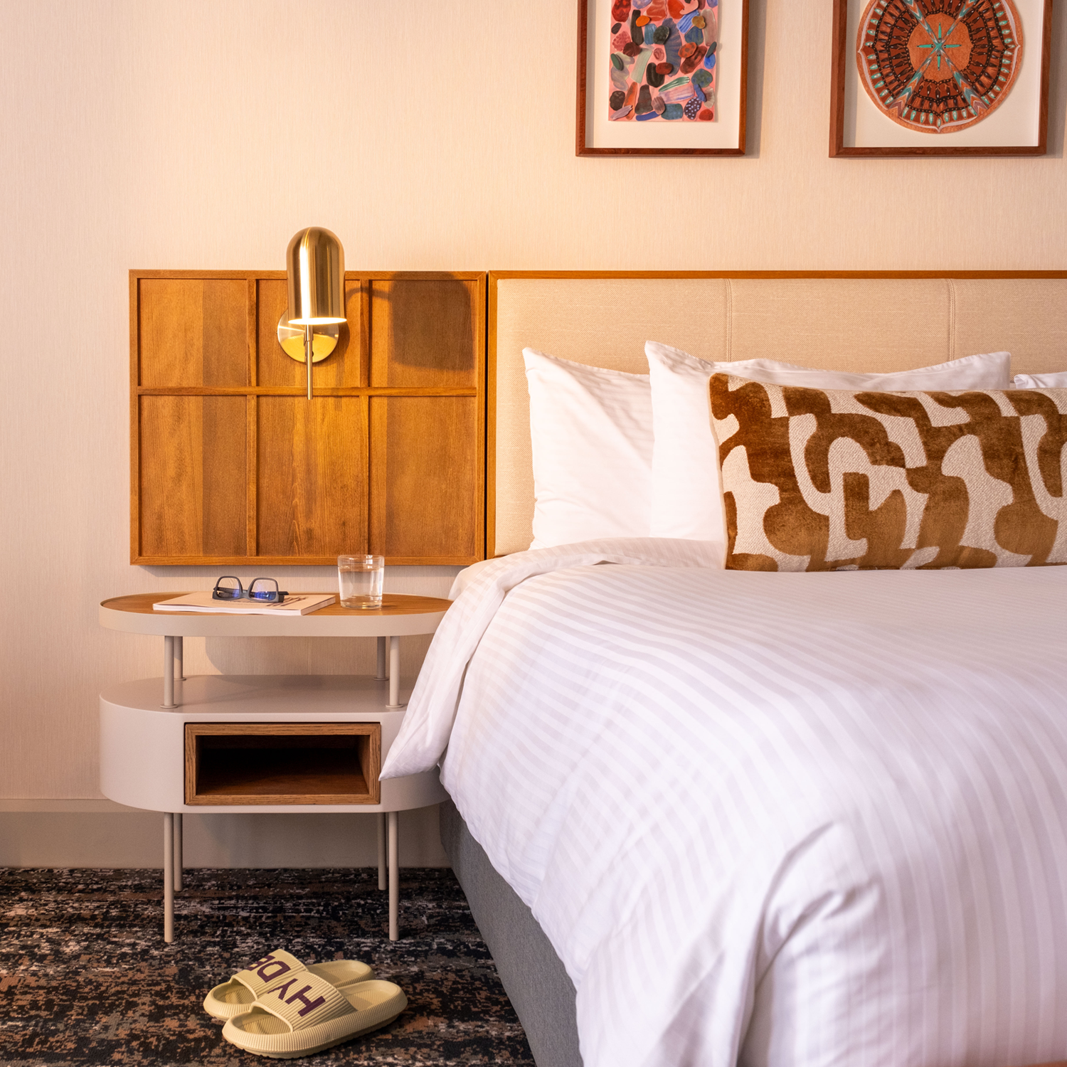 This is an image of a hotel room featuring a contemporary and stylish design. The room has a large bed with a tufted leather bench at the foot. The walls are decorated with soft pink tones and framed artwork above the headboard. The room includes a large, illuminated mirror with globe lights next to the bed, a wall-mounted TV, and a side table with a modern lamp and outlets. The window has sheer curtains, allowing natural light to filter in, and the ceiling shows an exposed concrete design.