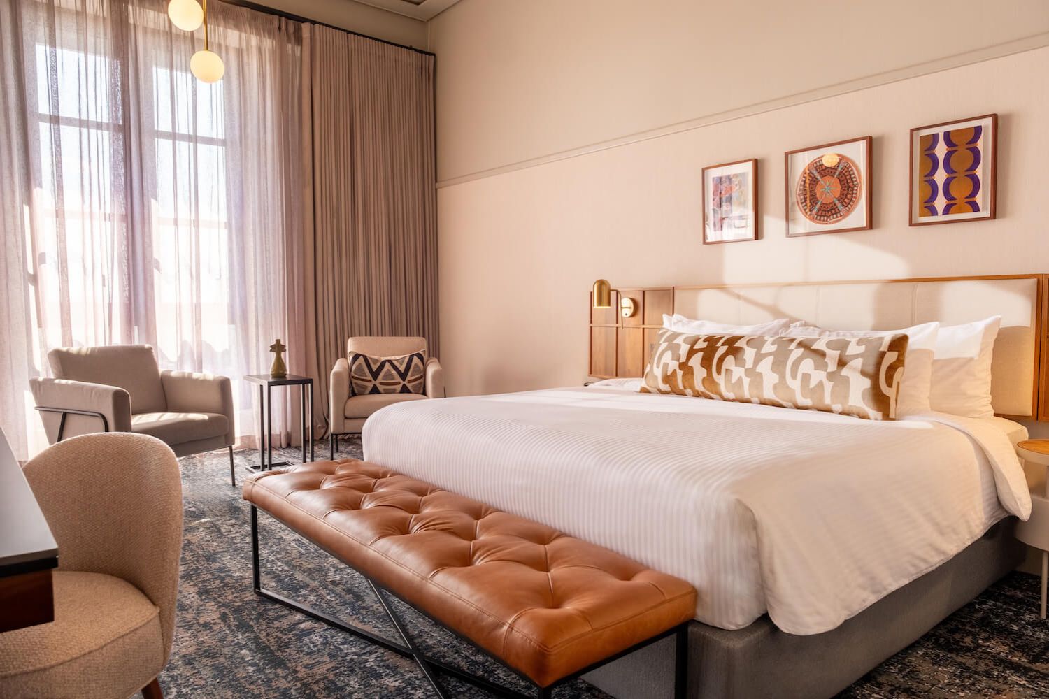 White bed angled with brown and beige headboard, brown leather bench in front of the bed, two armchairs with a small table between them, and white sheers covering large windows with tan curtains.