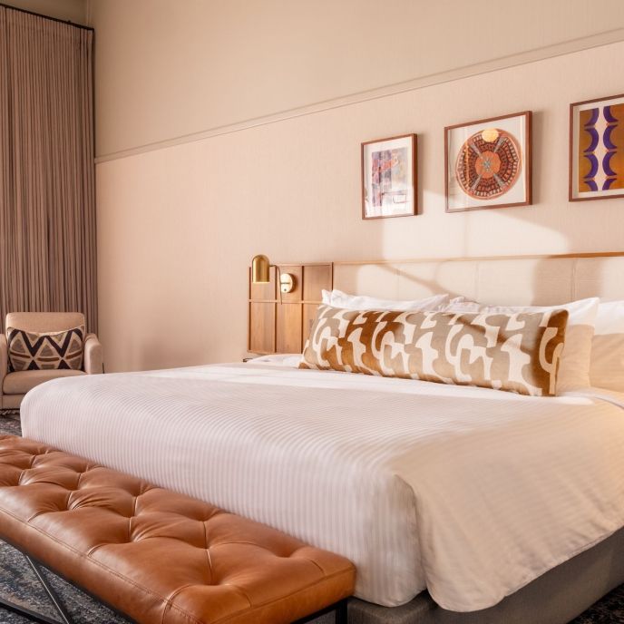 This is an image of a hotel room featuring a contemporary and stylish design. The room has a large bed with a tufted leather bench at the foot. The walls are decorated with soft pink tones and framed artwork above the headboard. The room includes a large, illuminated mirror with globe lights next to the bed, a wall-mounted TV, and a side table with a modern lamp and outlets. The window has sheer curtains, allowing natural light to filter in, and the ceiling shows an exposed concrete design.