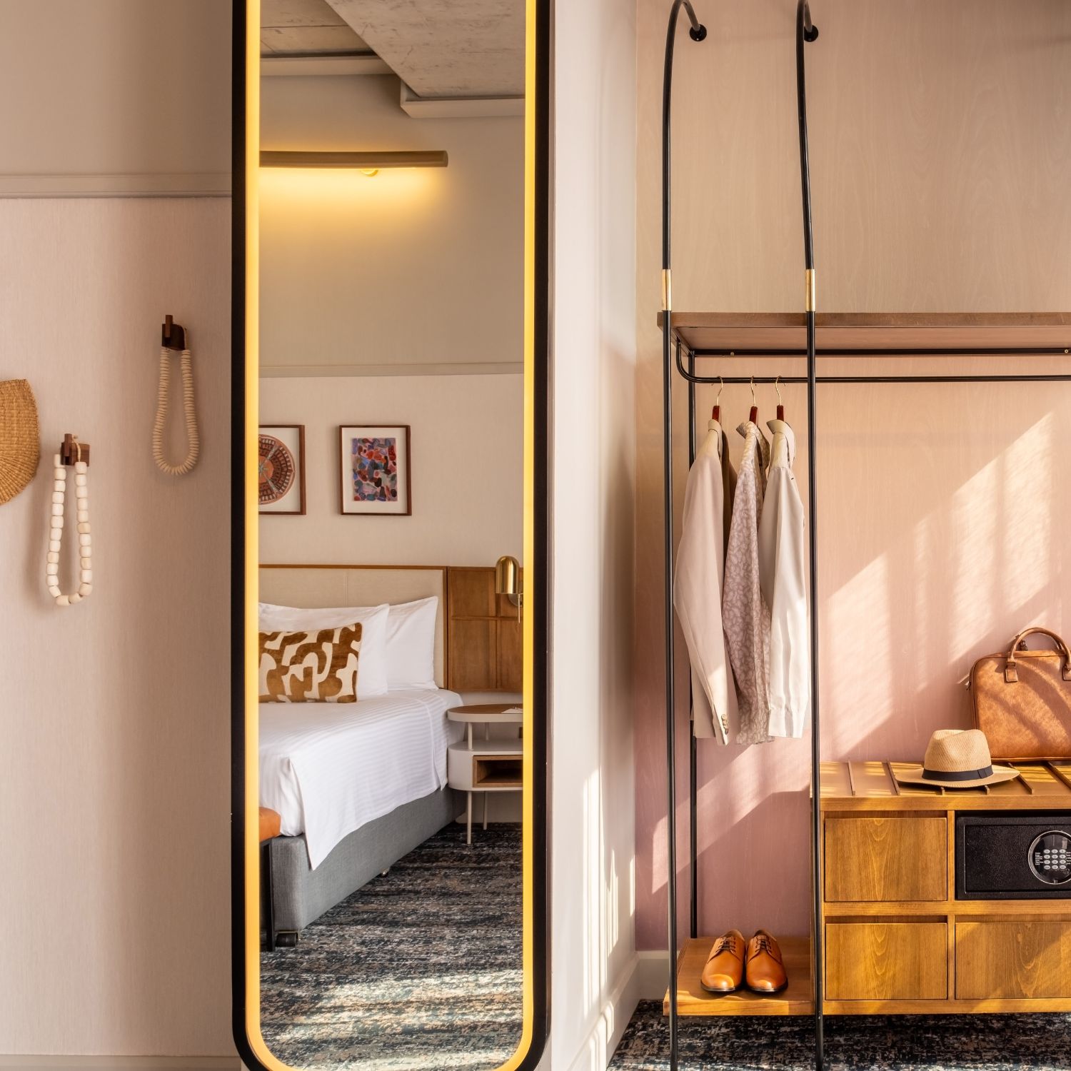 This is an image of a hotel room featuring a contemporary and stylish design. The room has a large bed with a tufted leather bench at the foot. The walls are decorated with soft pink tones and framed artwork above the headboard. The room includes a large, illuminated mirror with globe lights next to the bed, a wall-mounted TV, and a side table with a modern lamp and outlets. The window has sheer curtains, allowing natural light to filter in, and the ceiling shows an exposed concrete design.