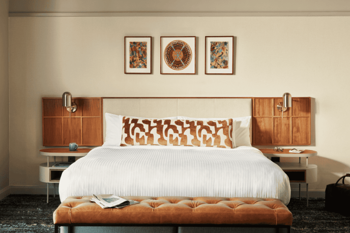 White bed with a brown and white scatter cushion, brown headboard with beige, bedside tables on either side of the bed and a tufted leather bench at the foot of the bed.